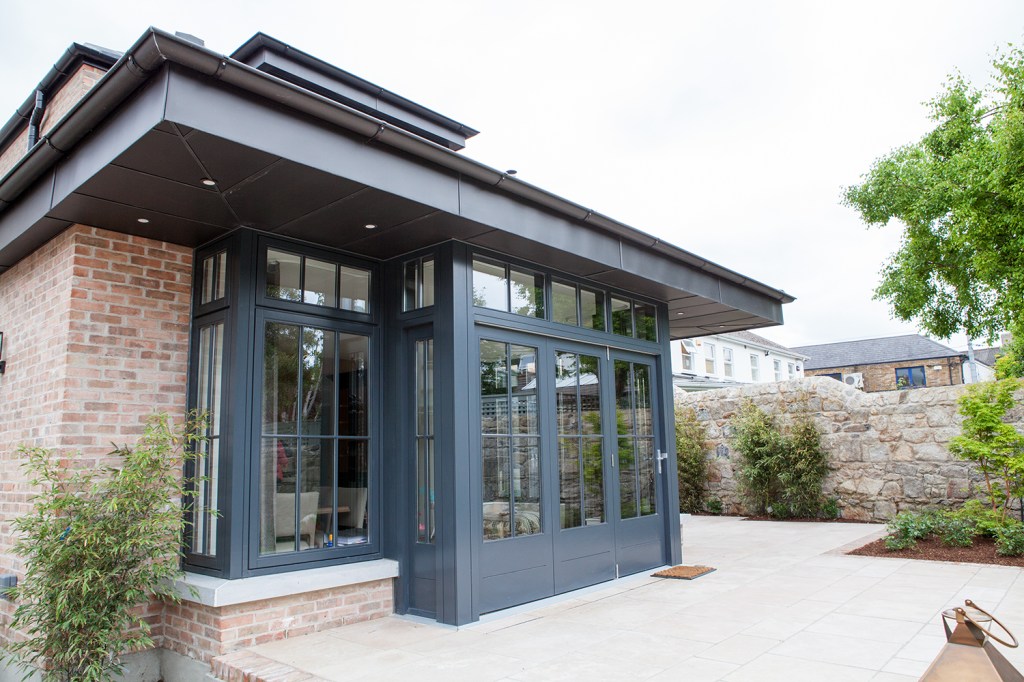 extended black roof with brick house - black wall windows with black frame and grille - fanlight windows - paving in front of house