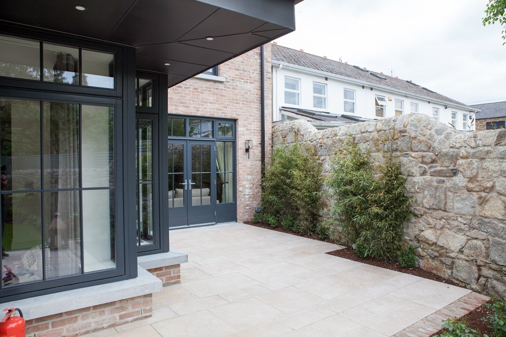 stone wall - black window frame and grille - wall windows - front yard paving - brick house - fanlight windows - extended roof