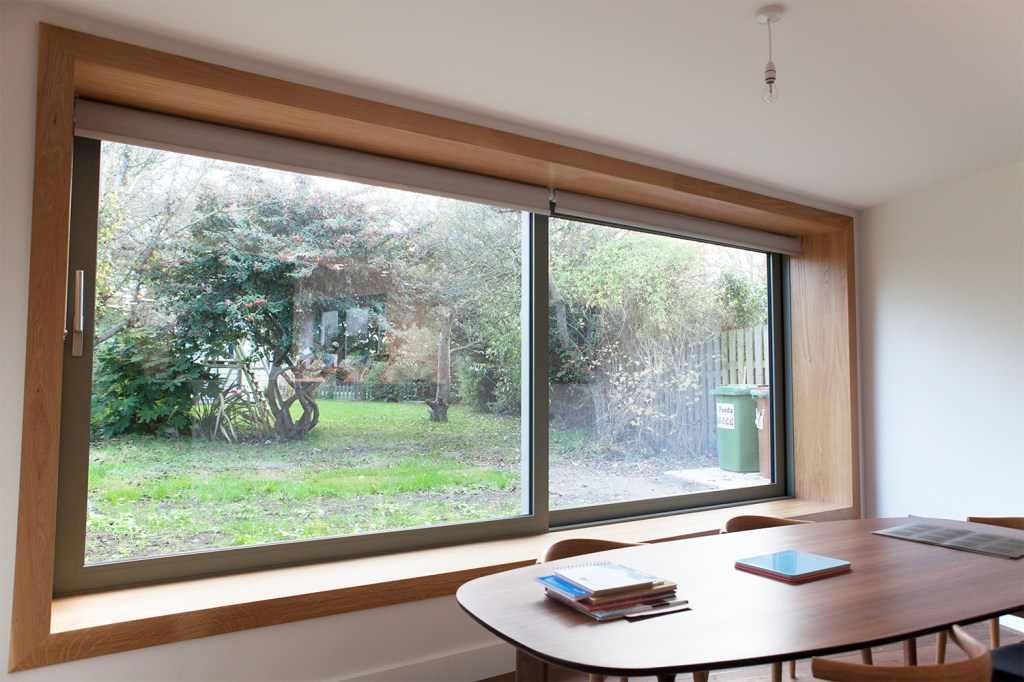 Big double clear sliding windows with green frame - wooden window casing with window sill - dining table - view of back garden with bush tree's