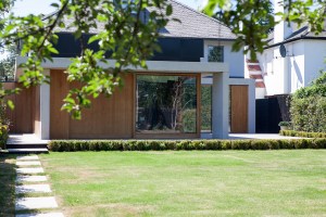 View through tree branches - wood sheeted walls with window walls and cement pillar - green grass landscape - pathway
