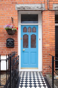 Walkway view of blue front door - transom door - door with stained glass windows - black iron baluster - checkered floor tile - pot plant with flowers hanging at front door