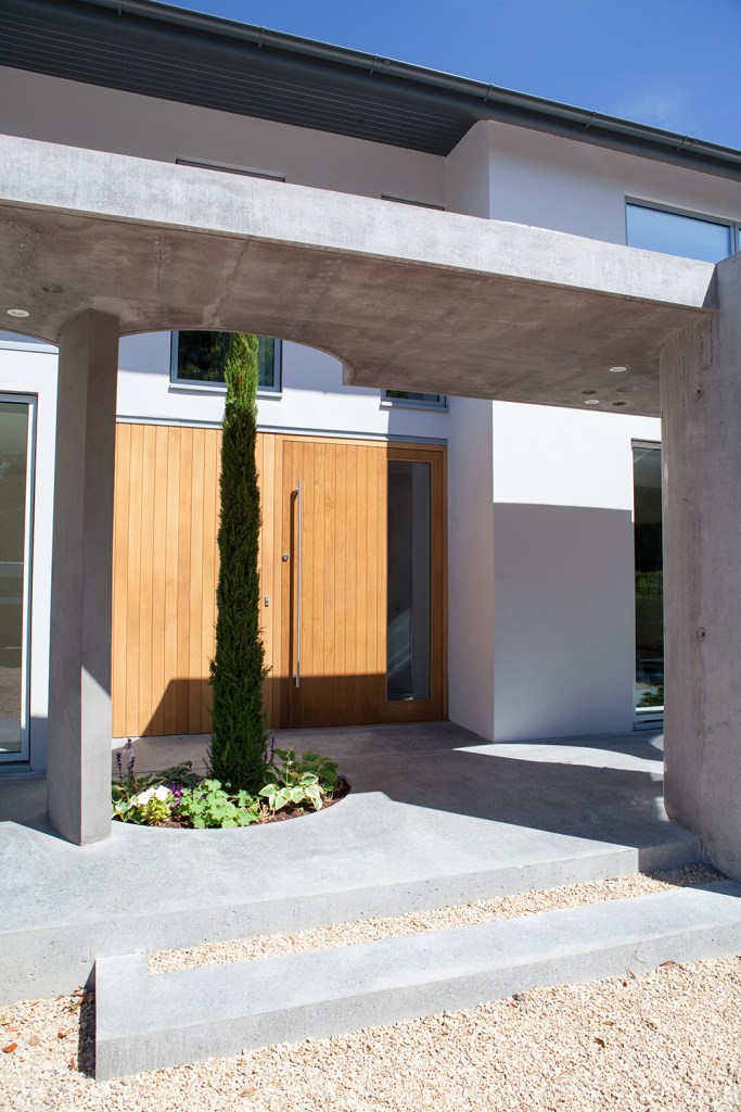 View to front door with cement pillars and patio - green thin tall tree - wooden double door with sidelite window - gravel