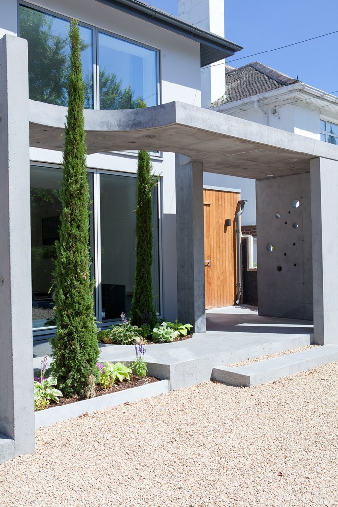 Cement pillars and extended roof on patio - thin green tree's in front - window walls - gravel