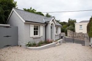 Little grey house with white double slide window with white window frames and white shutters - grey gate with black hinges - gravel
