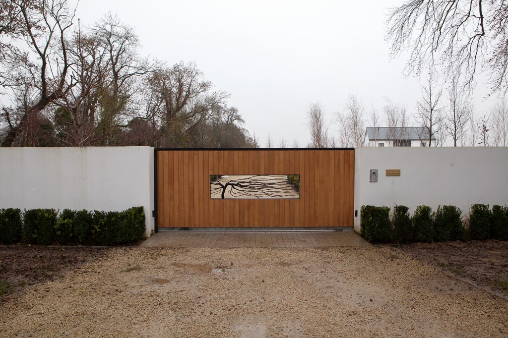Sheeted gate with metal tree inside gate - white walls - bush hedges - gravel road - tree's in background