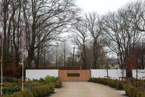 Photo of driveway gravel leading to sheeted gate - tree's in back ground - hedges on the side of driveway - white wall