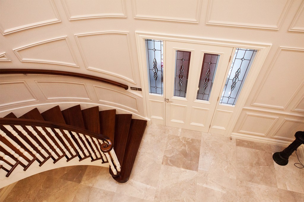 View of white front door with transom windows and white sidelite windows with black grill details - dark wood staircase with white baluster and dark wood staircase handrail - white panel walls and tile floor