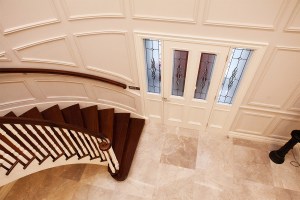 View of white front door with transom windows and white sidelite windows with black grill details - dark wood staircase with white baluster and dark wood staircase handrail - white panel walls and tile floor