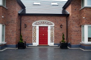 Front view of red front door with gold door knob and knocker - white transom windows with white sidelite windows and grills - two black pots with green small tree's - red brick house - black roof - tile front yard