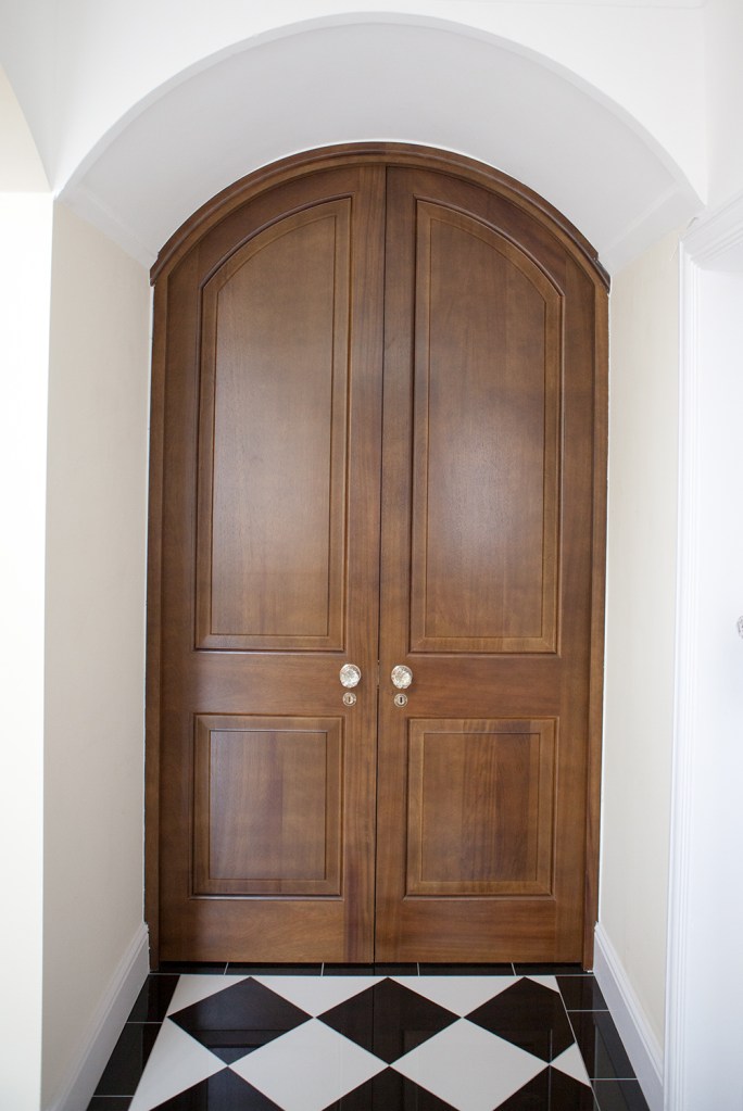 Arched wooden double side by side door - arched wood door frame - gold door knobs - white door casing - black and white tiled floor