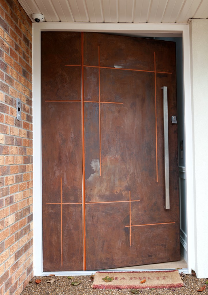 Big carved wood door with long silver handle - white door frame - doormat - brick wall - extended white patio roof