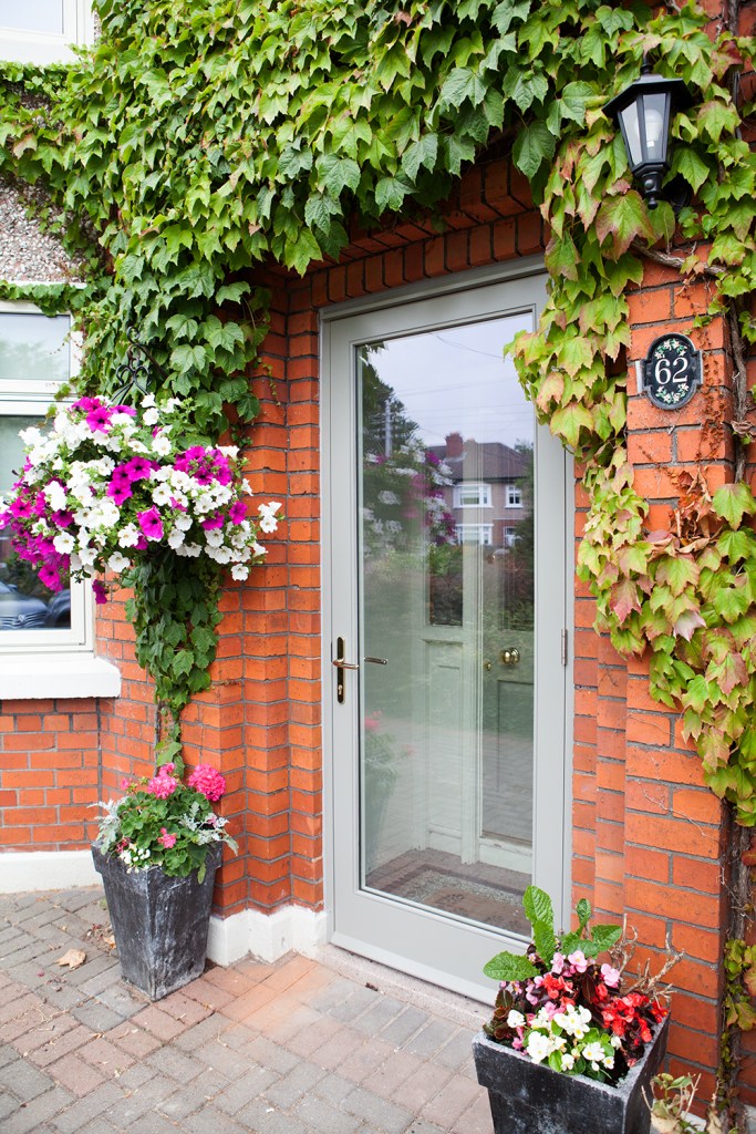 Transom door - grey door frame - vines on brick wall - red brick - pink and white flowers - black lantern light - pot plants - front door