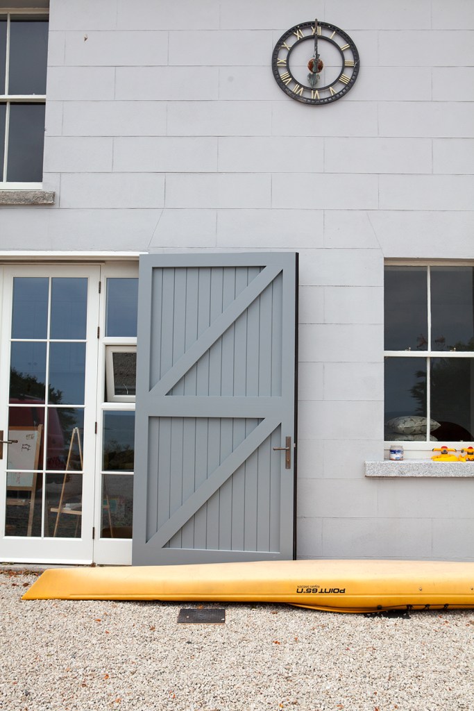 black outside clock on wall - yellow kayak - transom door - big grey outside door - grey cement block house - gravel