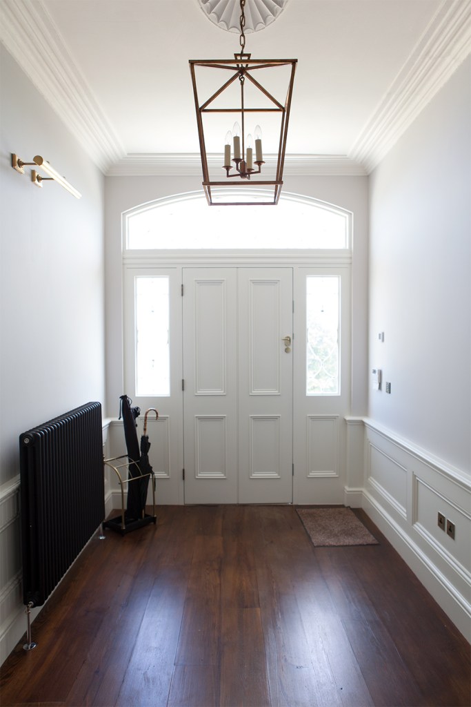 White entrance hall with white door and windows at the side and at the top of door - light hanging from roof - dark wood floors - black heater against wall