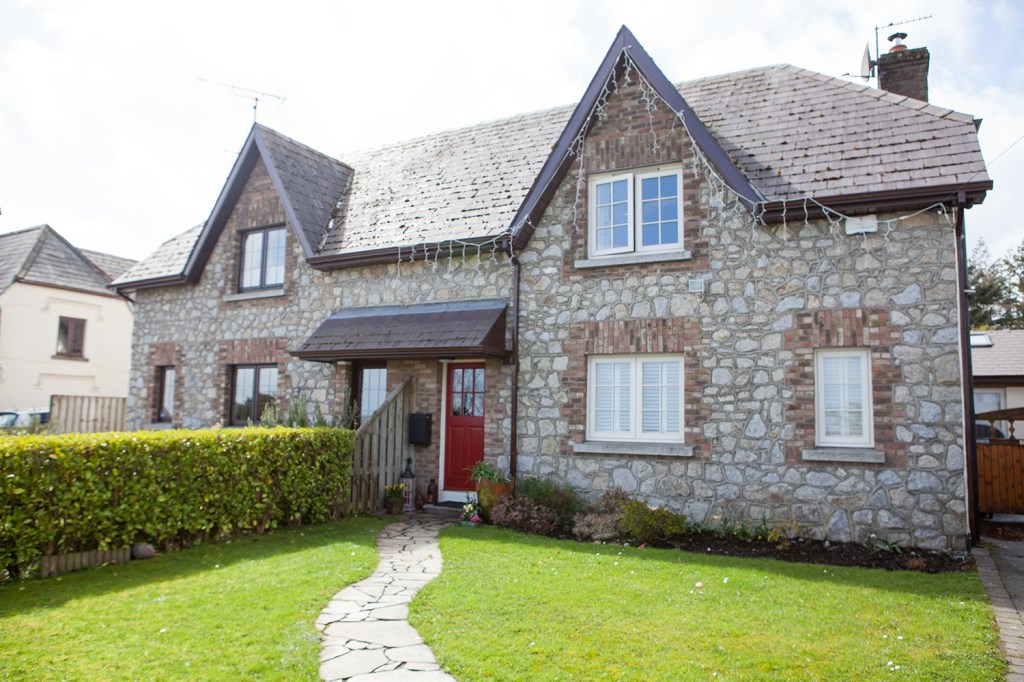 stone pathway leading to red door with stone house - green hedge and grass landscape - fairy lights hanging from roof - white framed windows with grilles