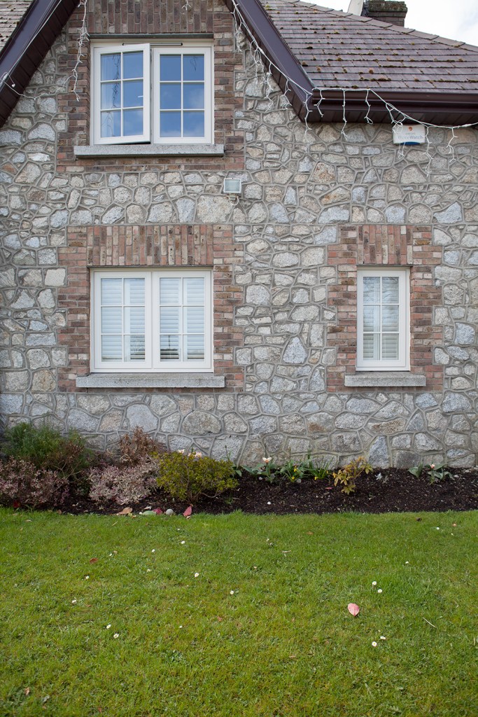 hanging fairy lights from roof gutter - white frame windows with grilles - windows with cement apron - bricks around window and stone walls - garden bed - grass lawn
