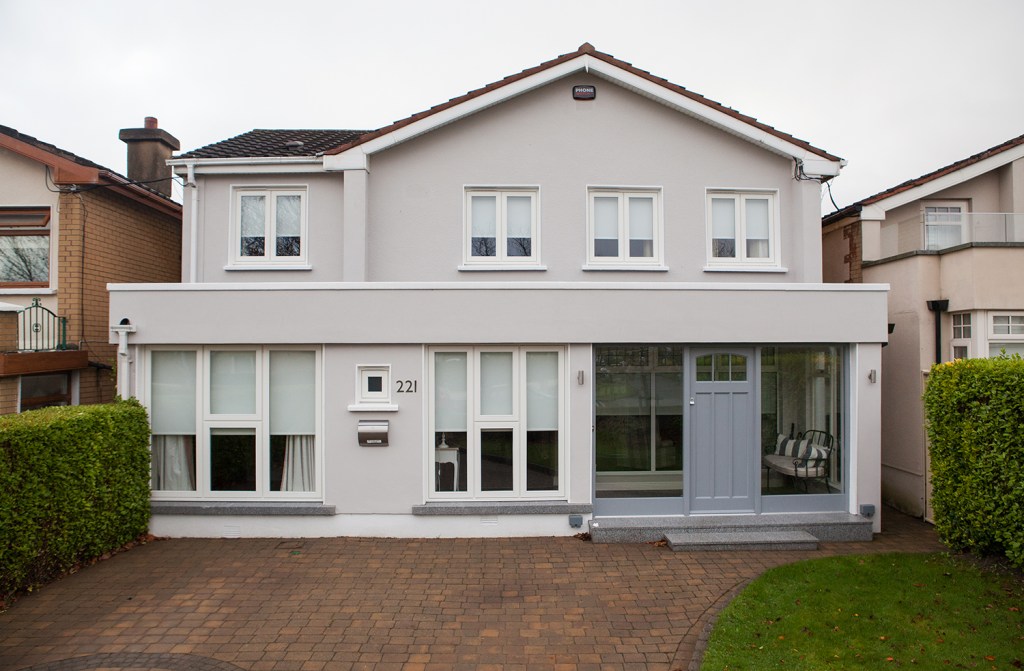 Brick driveway - bush hedges and small patch of grass - house with transom door and glass side windows - tall white framed windows on floor level postbox in wall