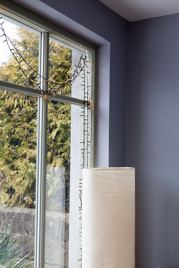 fairy lights hanging in window - tall lamp shade - grey wall corner - view of a tree - window with grilles and glass panes