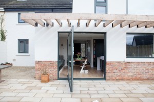 Open pivot door with black frame - glass pivot door - wooden beam roof extender - half way brick wall - outside view - white house