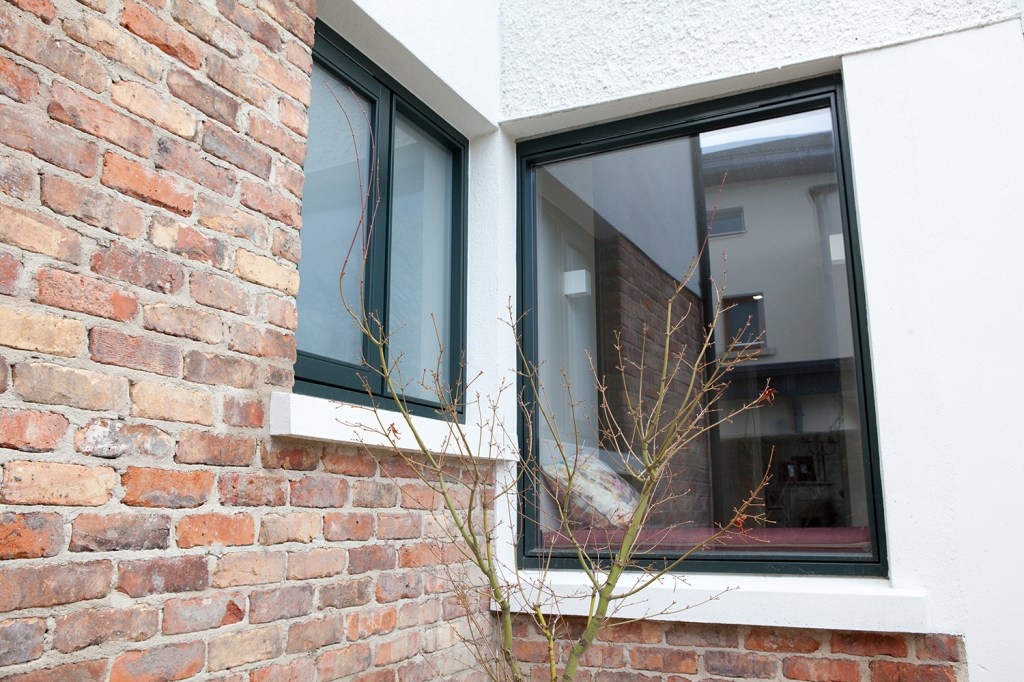 Brick house wall - white house wall - frosted window with black window frame and white arch - big clear window with black frame - small tree branches growing in front of windows