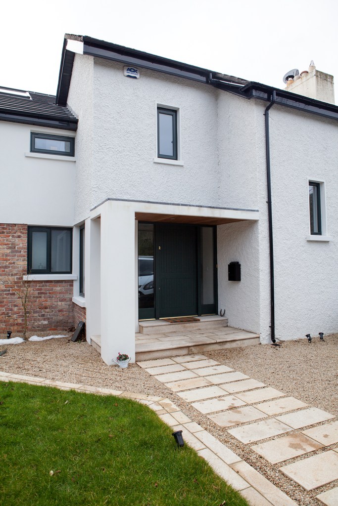 View of front of house - pathway to front patio and door - door with sidelites - half brick wall - green grass landscape - light grey house with black roof gutters - gravel - big black door